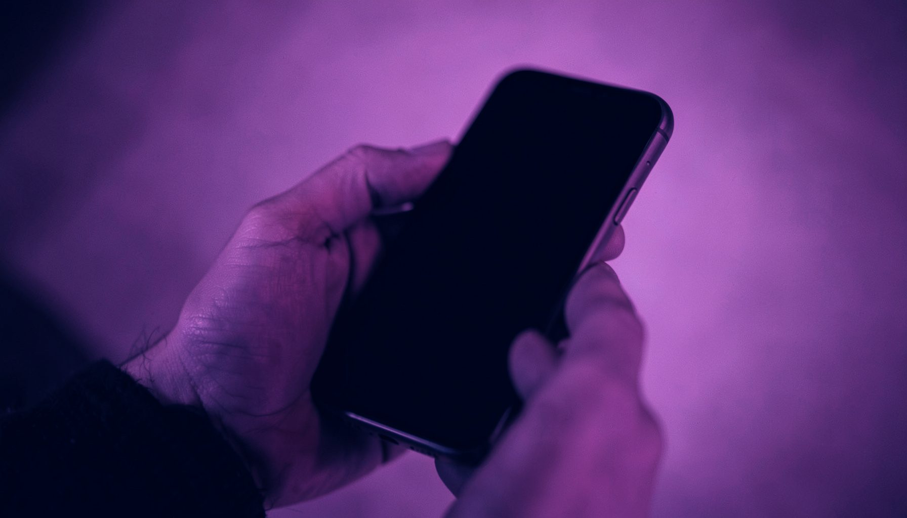 A moody duotone photograph in navy and magenta tones showing a person's hands holding a smartphone with a dark screen, set against a softly lit gradient background, evoking anticipation and modern mobile lifestyle.